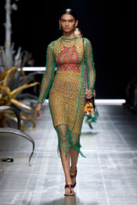 Runway model wearing a green mesh dress over an orange-red outfit, with gold hoop earrings and a brown handbag with tassels, walking the catwalk.