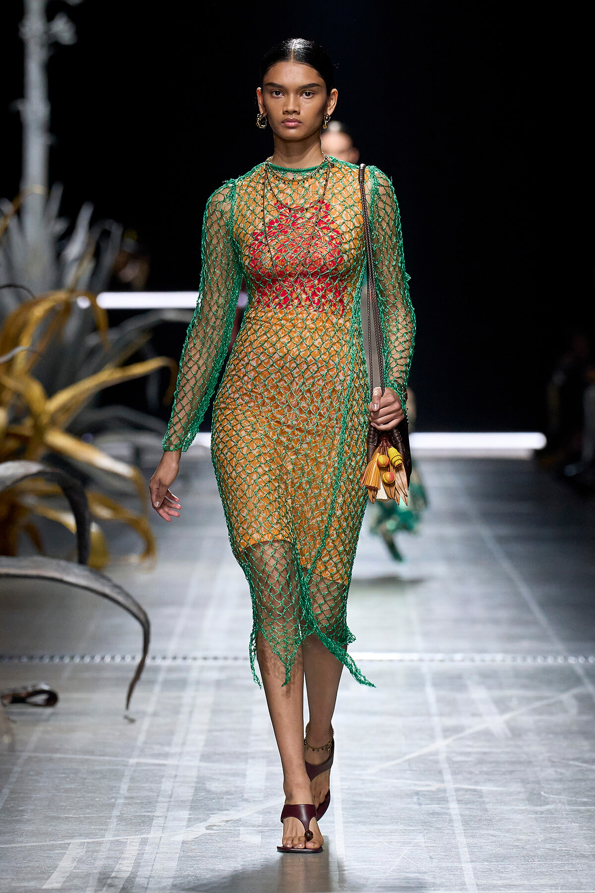 Runway model wearing a green mesh dress over an orange-red outfit, with gold hoop earrings and a brown handbag with tassels, walking the catwalk.