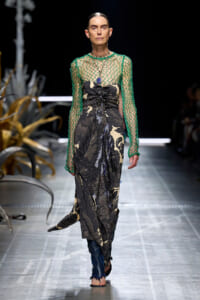 Model walking the runway in a sheer green mesh top over a black and beige floral-patterned dress with draped details and blue sandals.