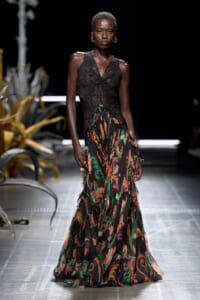 Model walking the runway in a black lace bodice and a long sheer floral-print skirt on a fashion show stage.