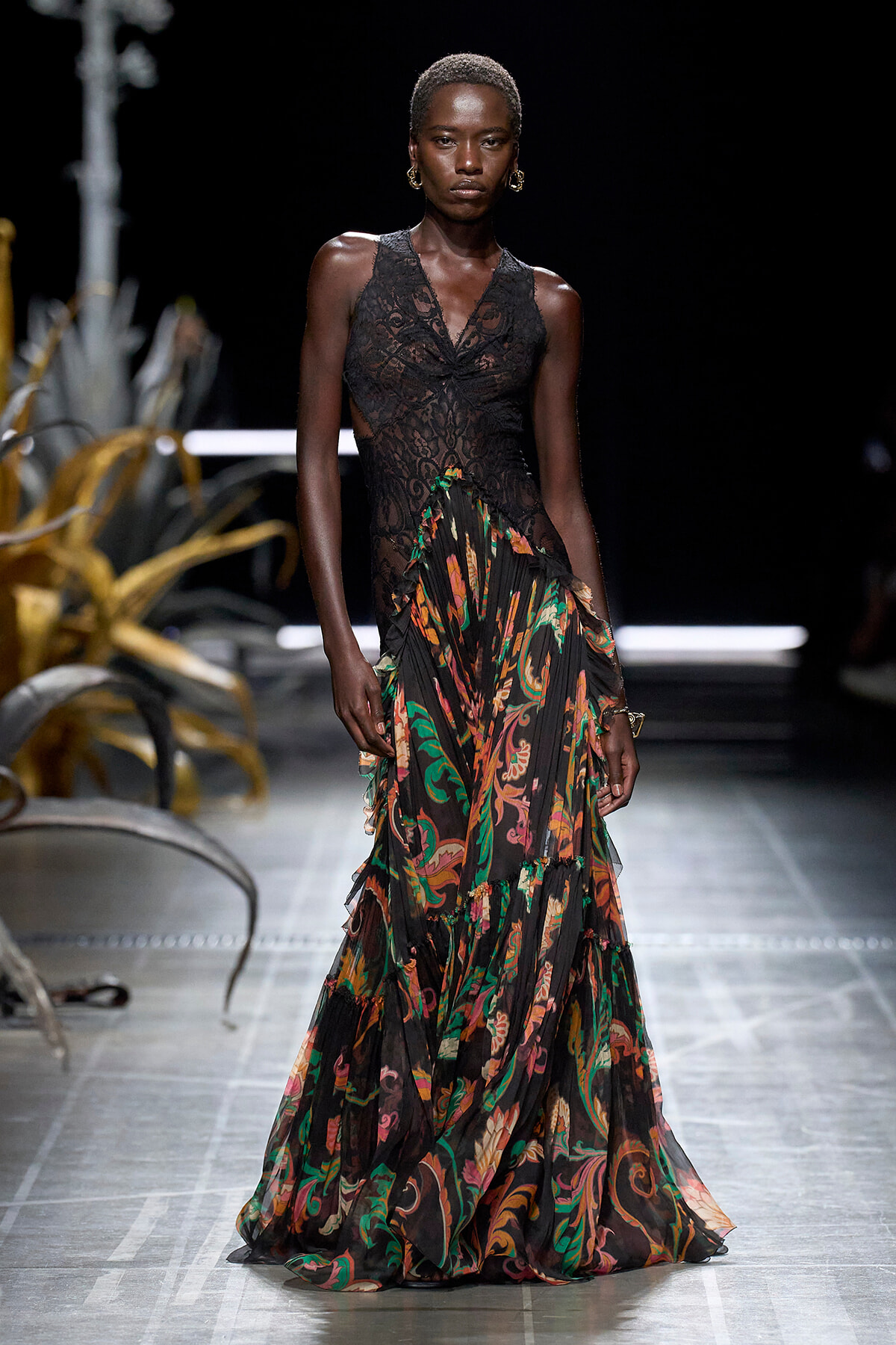 Model walking the runway in a black lace bodice and a long sheer floral-print skirt on a fashion show stage.