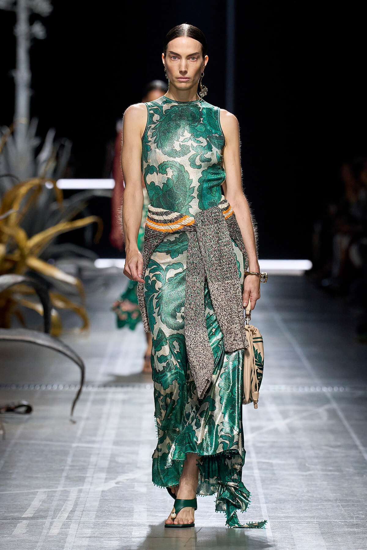 Model walking the runway in a sleeveless emerald-green botanical print dress with a textured scarf belt and beige handbag.