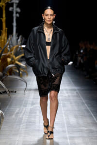 Model walking runway in a loose black quilted jacket over a sheer black lace skirt, layered jewelry accents her look.