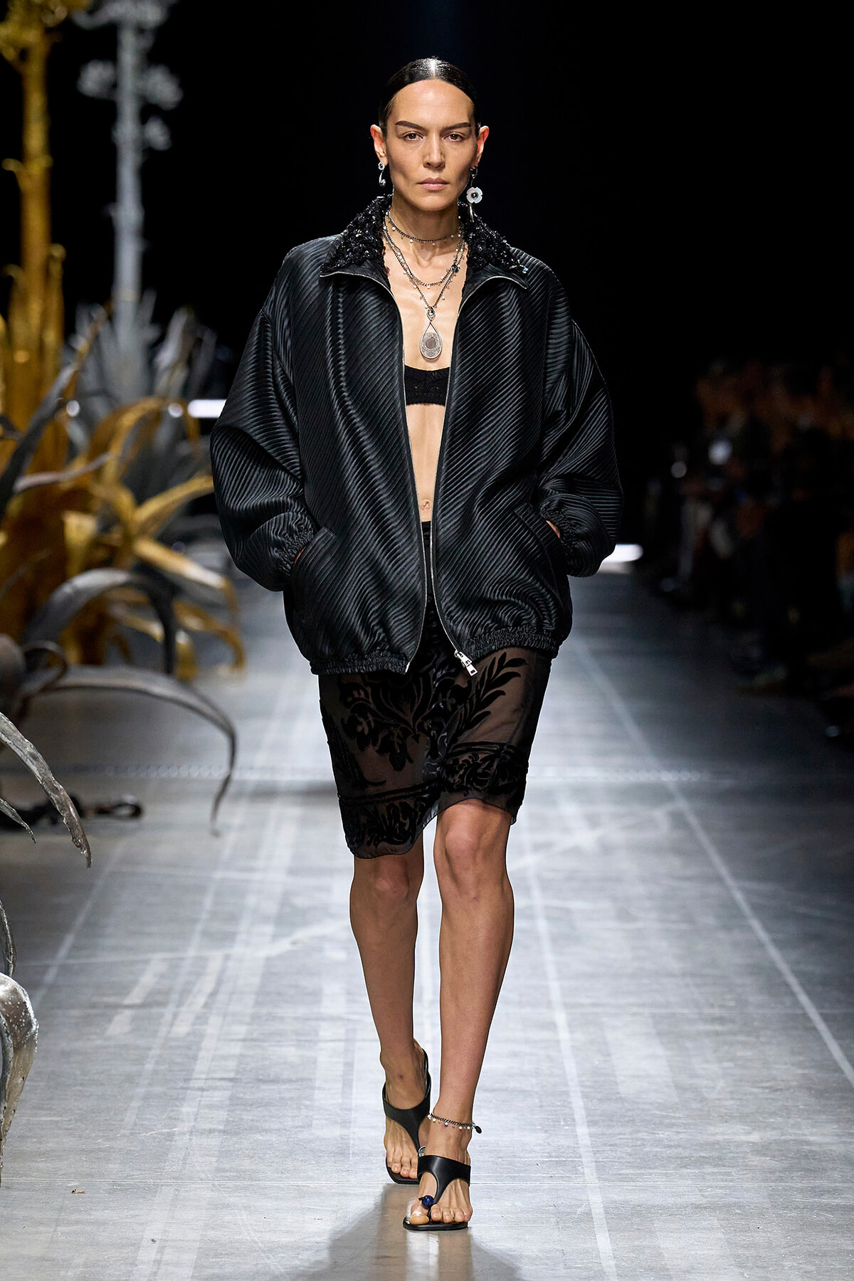 Model walking runway in a loose black quilted jacket over a sheer black lace skirt, layered jewelry accents her look.
