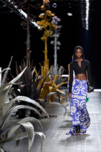 Model walks a fashion runway in a black hooded crop top and a blue-and-white ornate patterned maxi skirt, with large desert plants lining the catwalk.