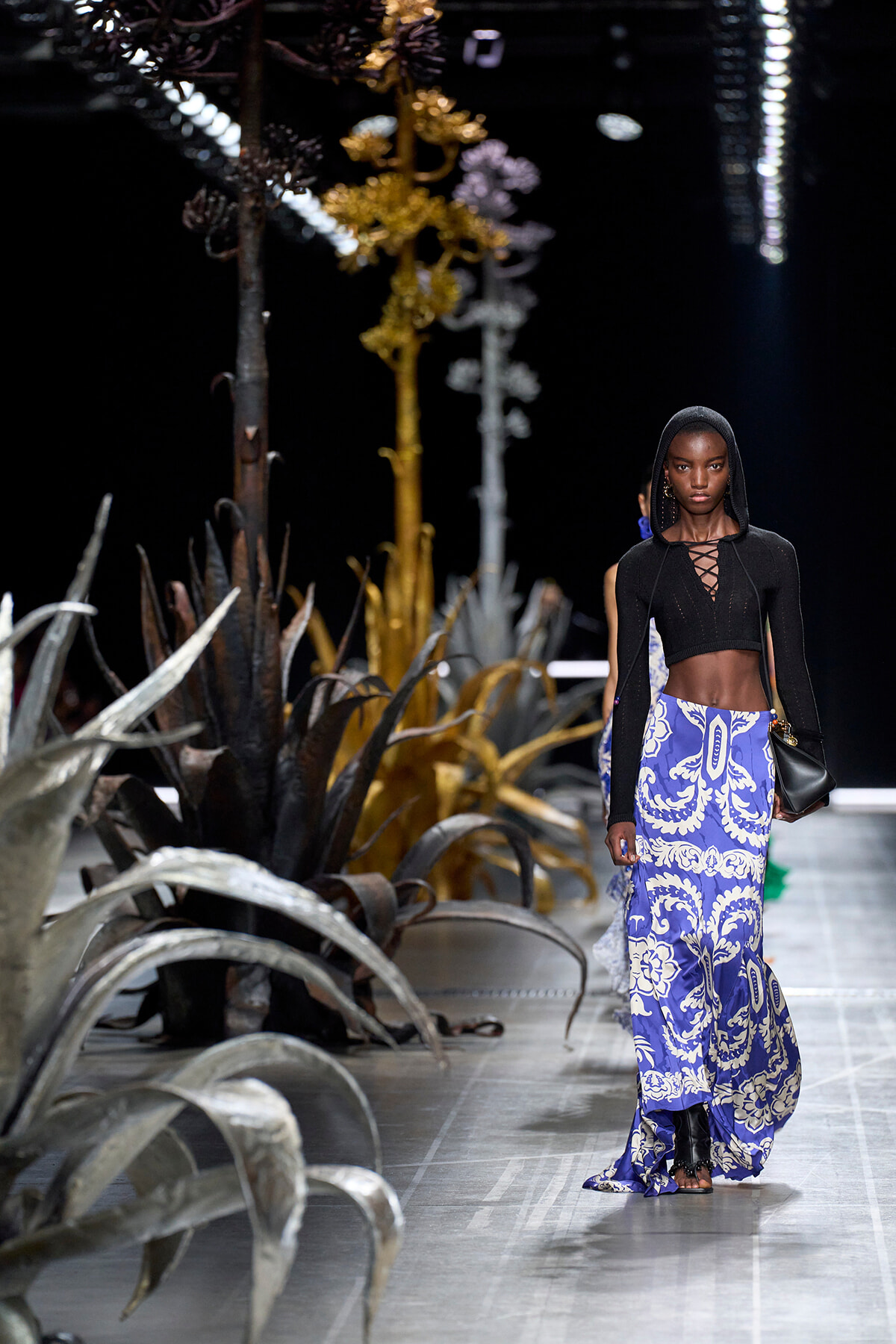 Model walks a fashion runway in a black hooded crop top and a blue-and-white ornate patterned maxi skirt, with large desert plants lining the catwalk.