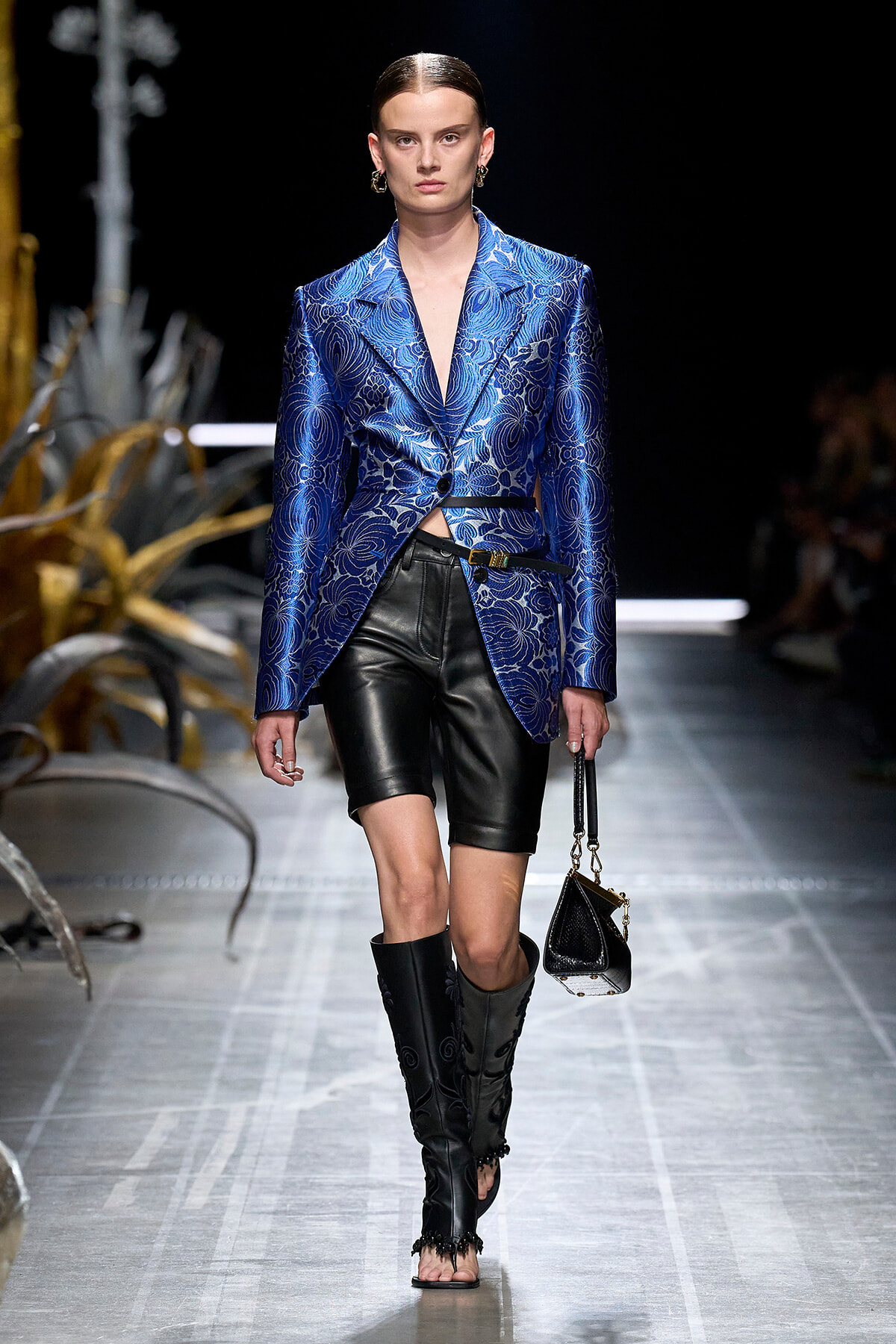 Model on a runway wearing a cobalt blue metallic blazer with a cropped waist, black leather shorts, knee-high boots, and a small black handbag.