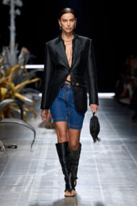 Runway model wearing a textured black blazer cinched at the waist, blue denim shorts, knee‑high black boots, and a small fringed black bag.