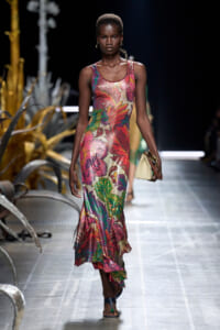 Model walking a runway in a colorful sleeveless floral maxi dress, carrying a beige handbag and wearing gold hoop earrings.