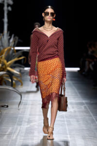 Model walking a fashion runway in a burgundy patterned knit top and orange fishnet skirt, with sunglasses and a brown handbag at her side.