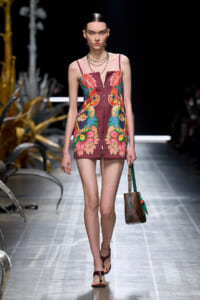 Model on a runway wearing a sleeveless maroon mini dress with bold tropical floral print and front zipper, paired with brown sandals and a brown bag with green strap.