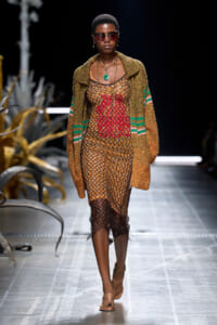 Runway model wearing a sheer lattice dress with a bold red floral design, overlaid by a chunky brown knit cardigan and green pendant necklace with sunglasses.",