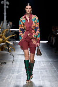 Model walks the runway in a burgundy floral-print shirt-dress paired with matching shorts, green embroidered boots, and a small brown handbag.