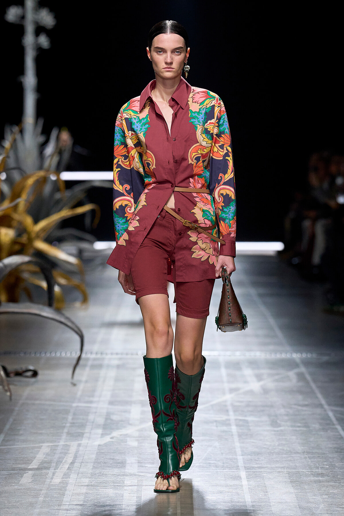 Model walks the runway in a burgundy floral-print shirt-dress paired with matching shorts, green embroidered boots, and a small brown handbag.