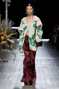 Model on a runway wearing a cream knit cardigan with green pattern over burgundy floral sheer pants, accessorized with green earrings and a burgundy shoulder bag.