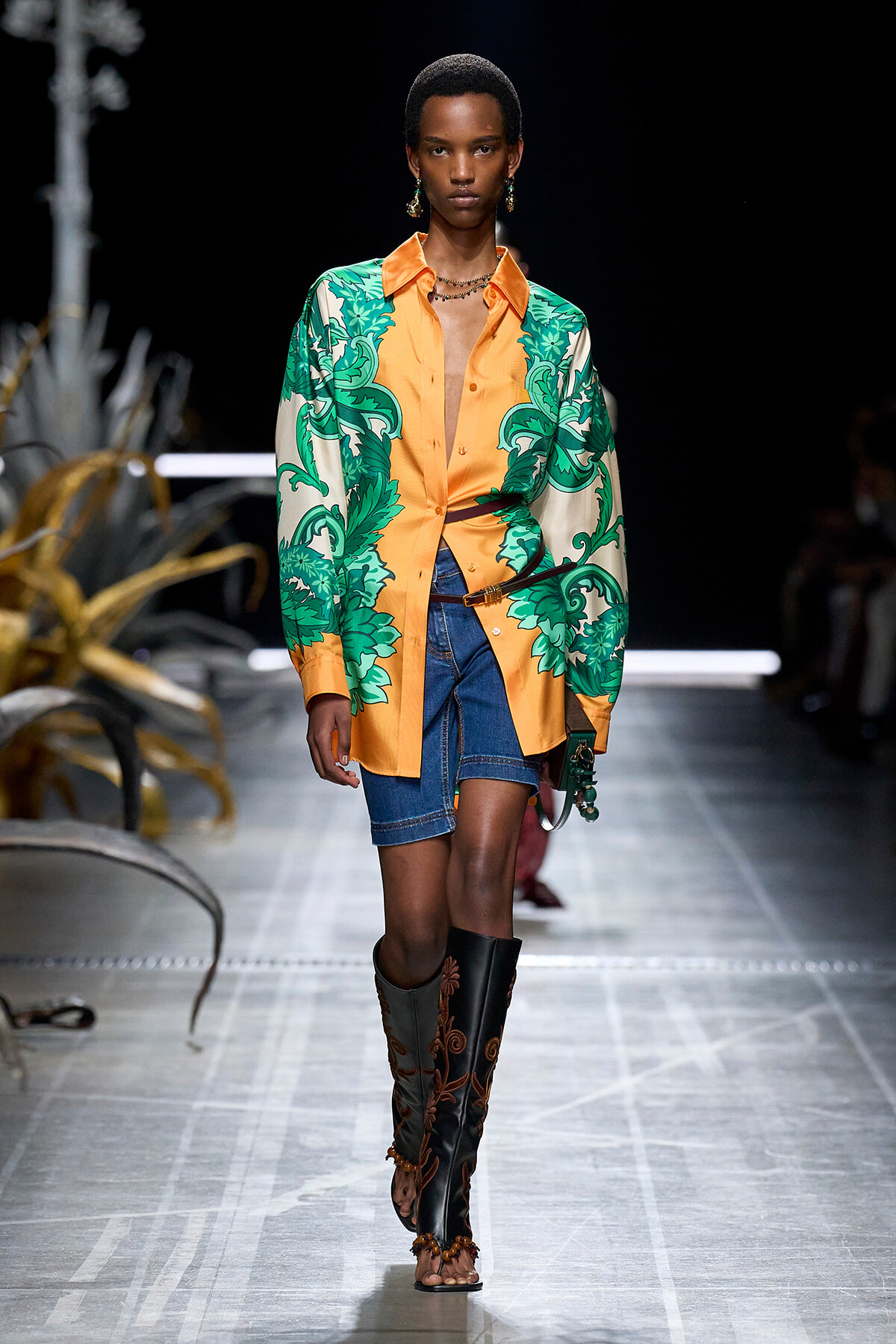 Model walking a runway in an oversized orange shirt with green botanical print over denim shorts and tall black embroidered boots.