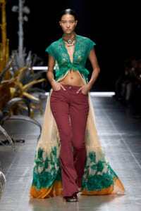 Model walks the runway in a teal lace crop top, open-front gown, and burgundy flared pants with a sheer, floral-printed train trailing behind.