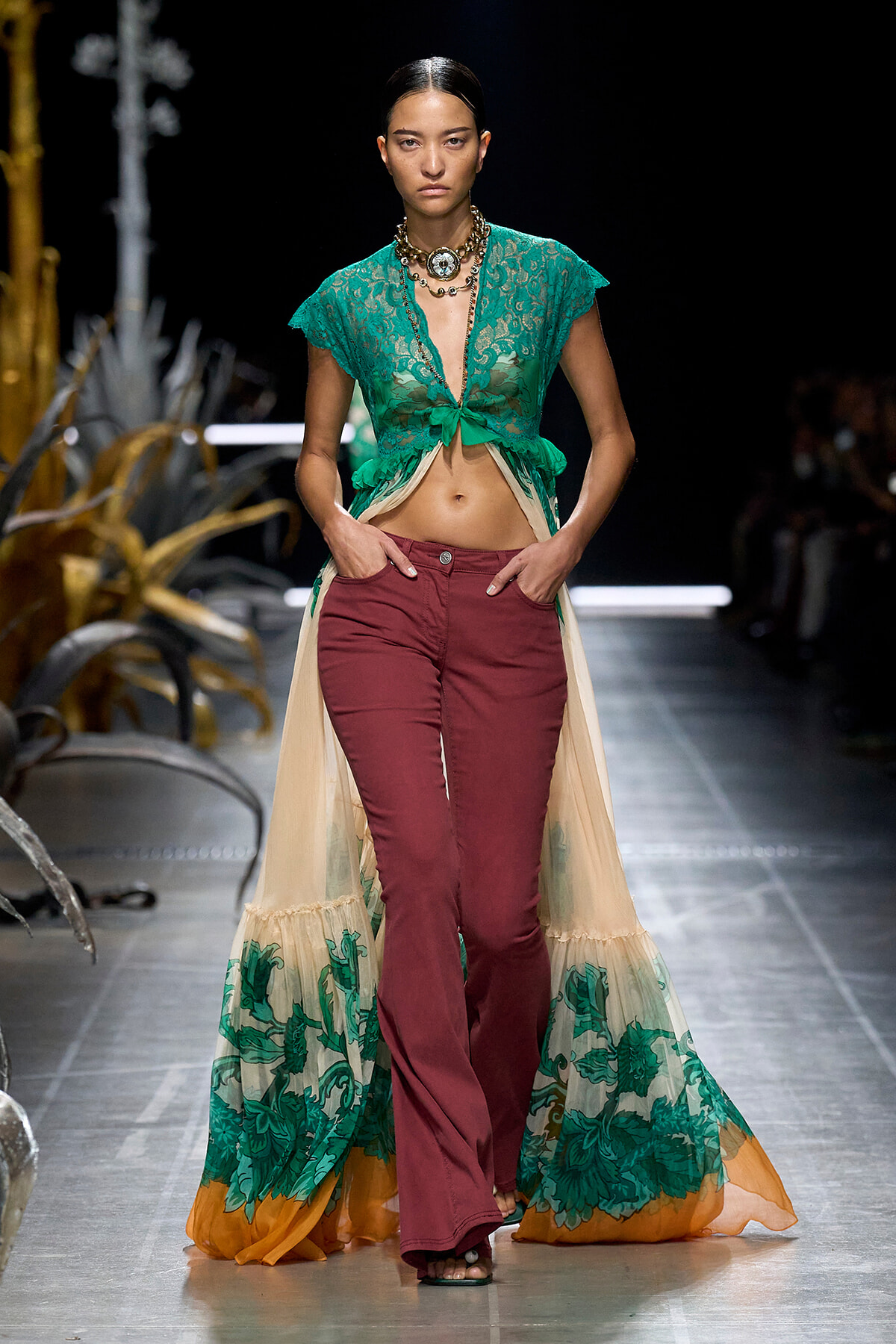 Model walks the runway in a teal lace crop top, open-front gown, and burgundy flared pants with a sheer, floral-printed train trailing behind.