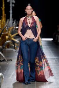 Runway model wears a sheer purple lace halter over jeans, accessorized with layered necklaces on a fashion show catwalk.