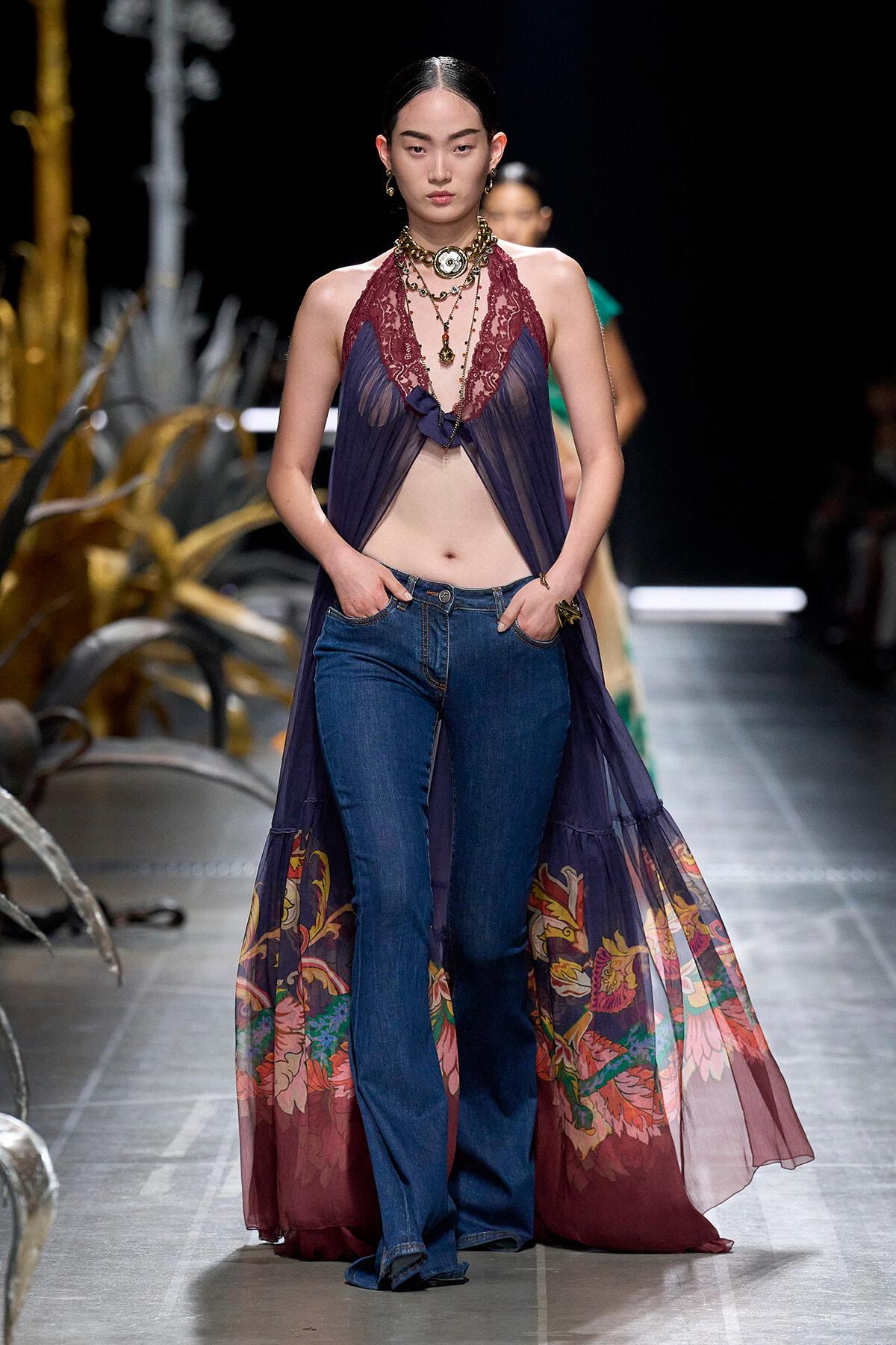 Runway model wears a sheer purple lace halter over jeans, accessorized with layered necklaces on a fashion show catwalk.