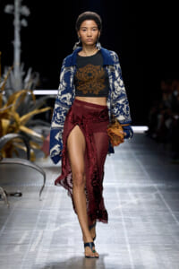 Model walks a fashion runway wearing a blue patterned cardigan over a black crop top with a brown floral design and burgundy lace-front skirt.