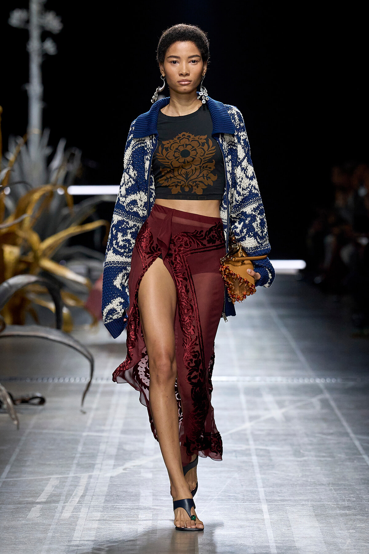 Model walks a fashion runway wearing a blue patterned cardigan over a black crop top with a brown floral design and burgundy lace-front skirt.