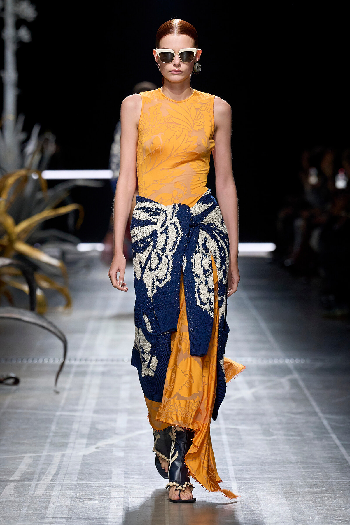 Model walks a runway in an orange sleeveless top with a botanical pattern, a navy floral knit wrap tied at the waist, and an orange patterned skirt, wearing white sunglasses.