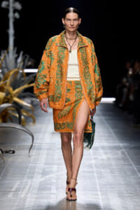 Model walks a runway in an orange-green sequined ensemble with a matching jacket and slit skirt, carrying a green bag.