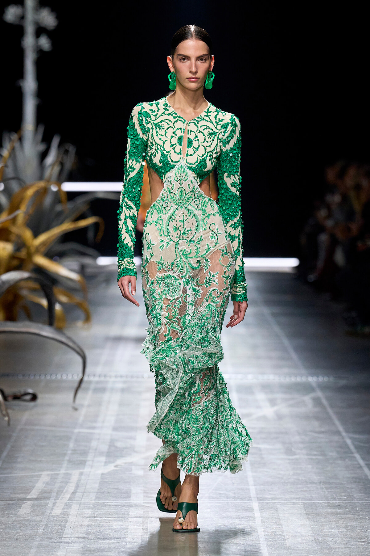 Model walks a runway in a green-and-white lace gown with floral embroidery, sheer panels, and cutouts at the waist, accessorized with green earrings and sandals.
