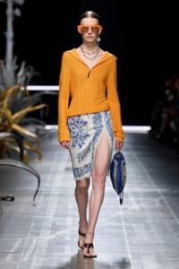 Model walks a fashion runway wearing an orange knit top with a blue lace-patterned skirt and a high slit, accessorized with orange sunglasses and a blue handbag.