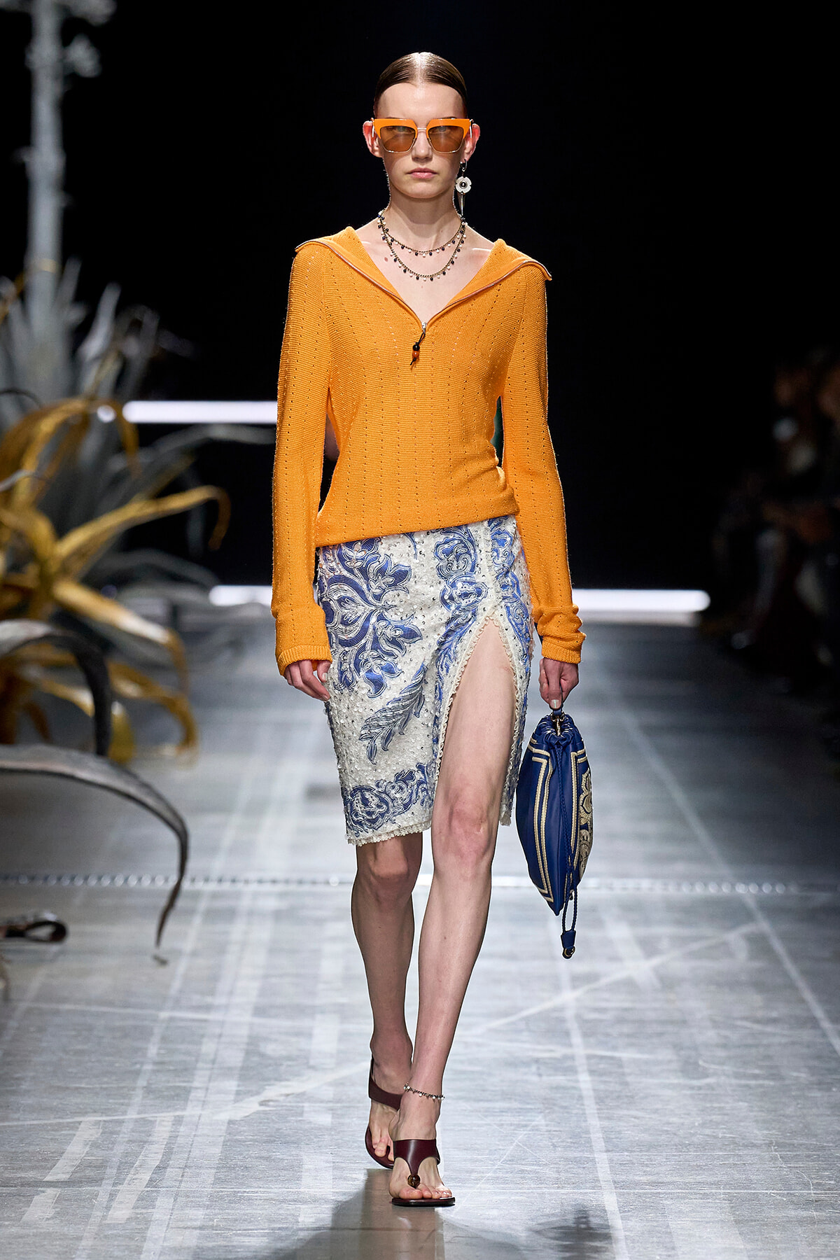Model walks a fashion runway wearing an orange knit top with a blue lace-patterned skirt and a high slit, accessorized with orange sunglasses and a blue handbag.