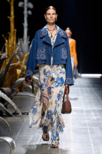 Model walks the runway in a blue denim jacket with geometric blue embroidery over a flowing blue-and-cream floral dress, carrying a brown studded handbag.