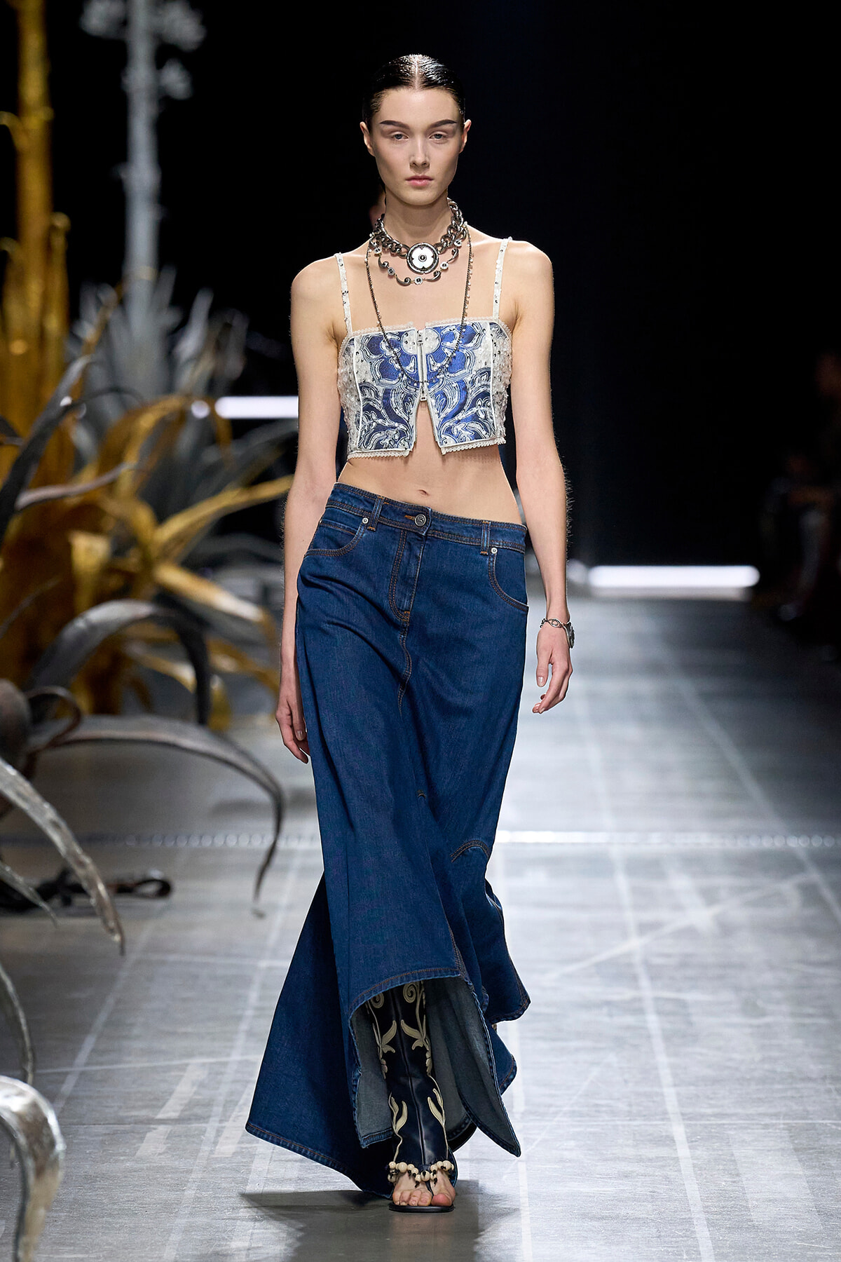 Model walks the runway in a blue patterned crop top paired with wide-leg denim jeans and layered necklaces on a dark catwalk edge.