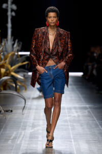 Model walks the runway in a red and black floral-patterned blazer over a plunging top, paired with blue denim shorts and black sandals with ankle jewelry.