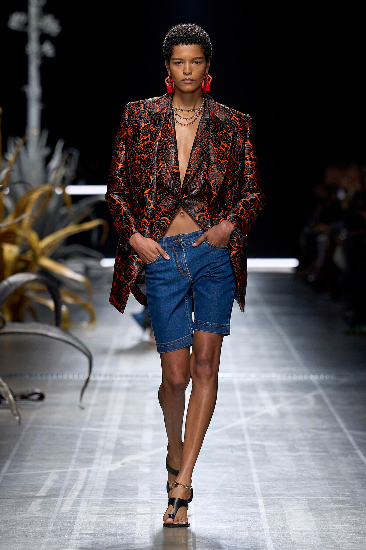 Model walks the runway in a red and black floral-patterned blazer over a plunging top, paired with blue denim shorts and black sandals with ankle jewelry.