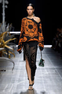 Model walks runway in an oversized orange and black floral knit sweater with one shoulder exposed, paired with a sheer black lace skirt and green-beaded handbag.