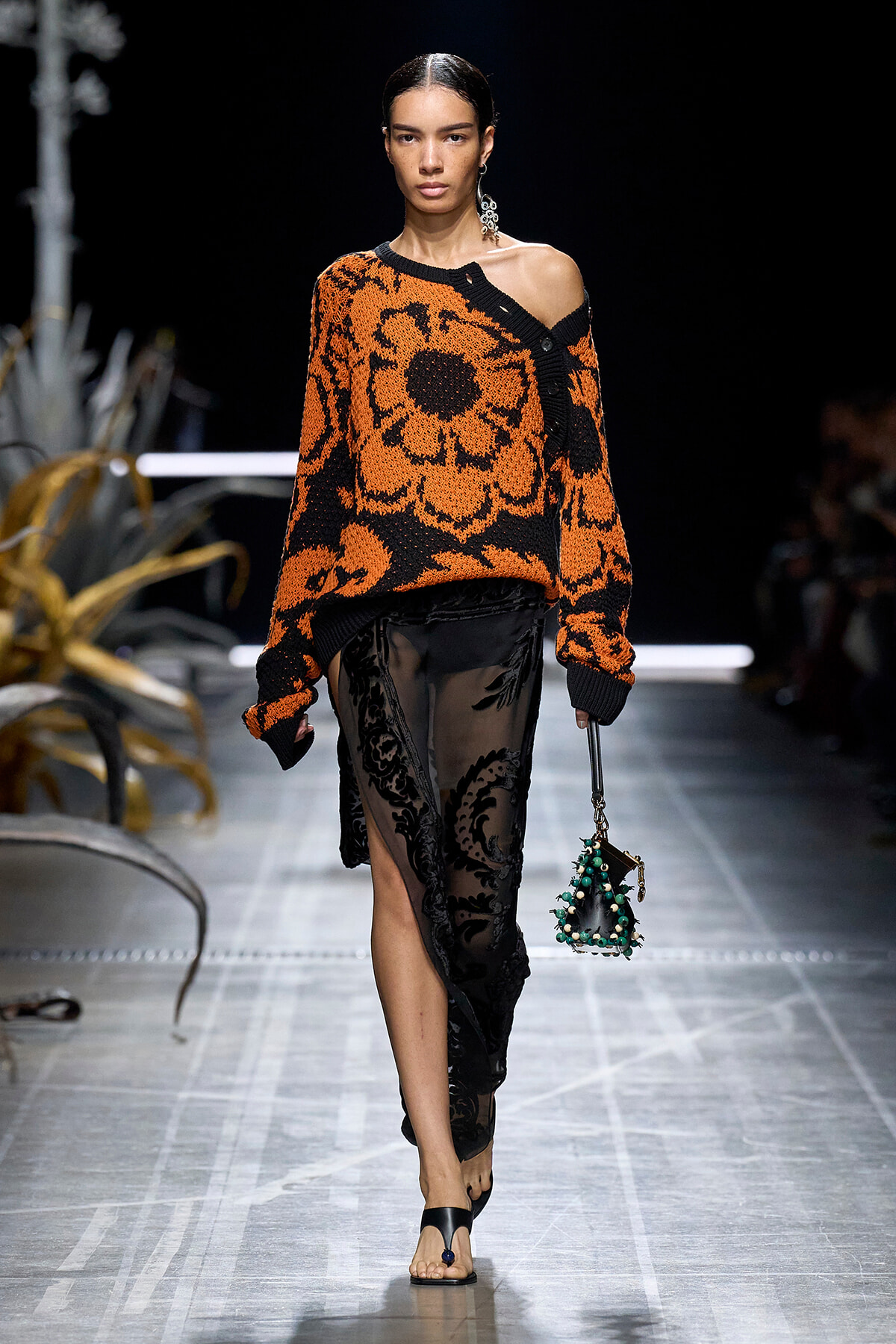 Model walks runway in an oversized orange and black floral knit sweater with one shoulder exposed, paired with a sheer black lace skirt and green-beaded handbag.