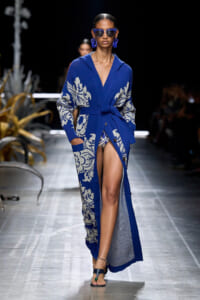 Female model walks the runway in a cobalt blue knit cardigan with a white floral pattern, belted at the waist, revealing a high slit and leg, paired with blue sunglasses and bold earrings.