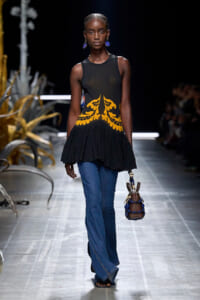 Model walks runway in a sleeveless black top with orange print, flared blue jeans, and a blue beaded handbag.