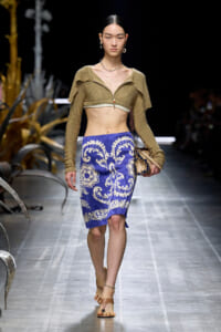 Model walking runway in a gold cropped knit top and blue white patterned knee-length skirt; beige sandals and accessories visible.