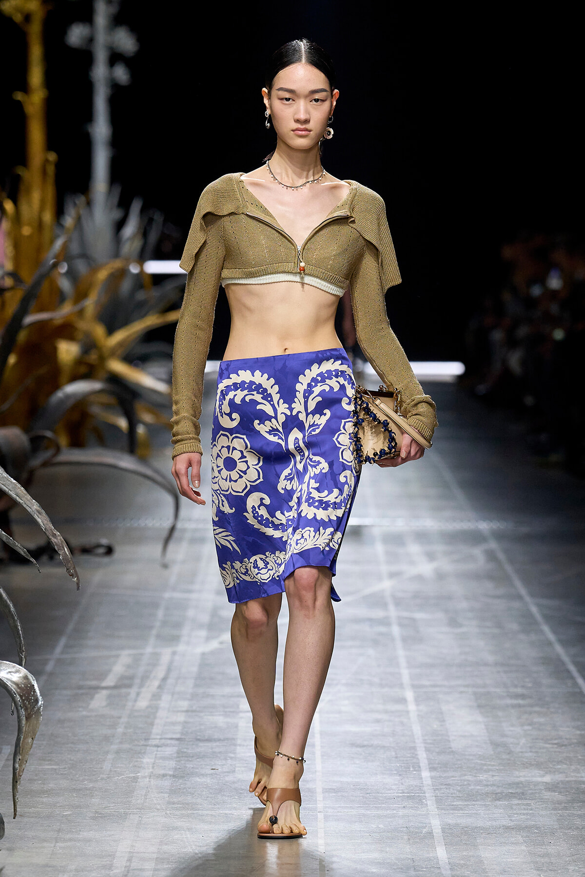 Model walking runway in a gold cropped knit top and blue white patterned knee-length skirt; beige sandals and accessories visible.