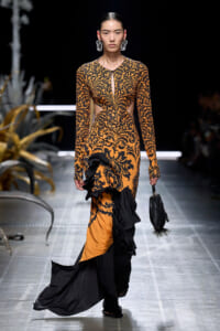Runway model in an orange and black damask-pattern gown with side cutouts and a ruffled black-and-orange train, carrying a black handbag.