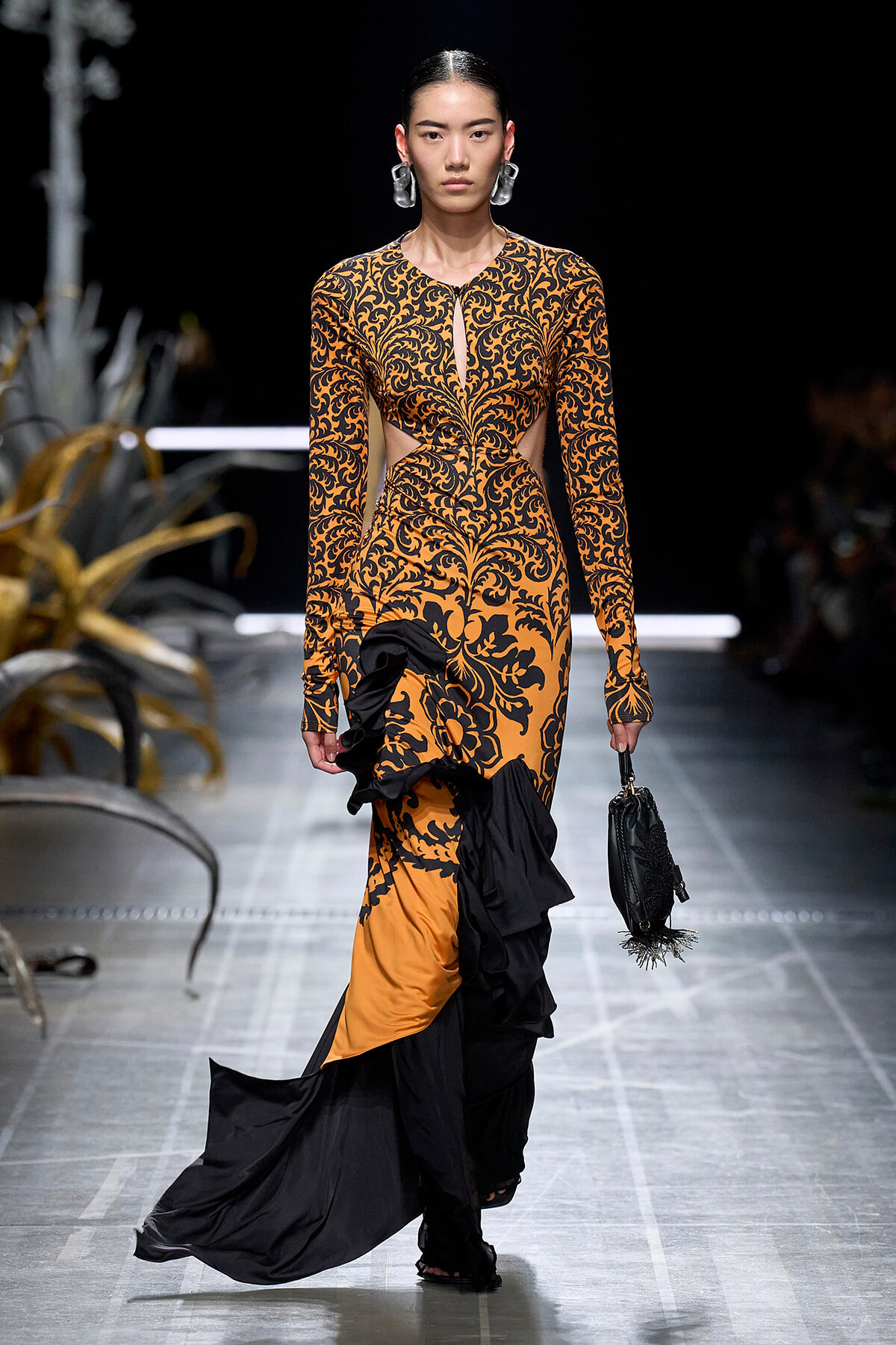Runway model in an orange and black damask-pattern gown with side cutouts and a ruffled black-and-orange train, carrying a black handbag.