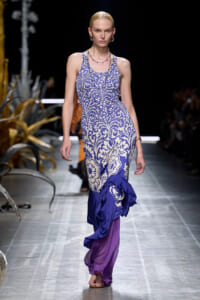 Model on a runway wearing a sleeveless blue and white damask dress with a purple ruffle hem and layered fabric at the bottom.