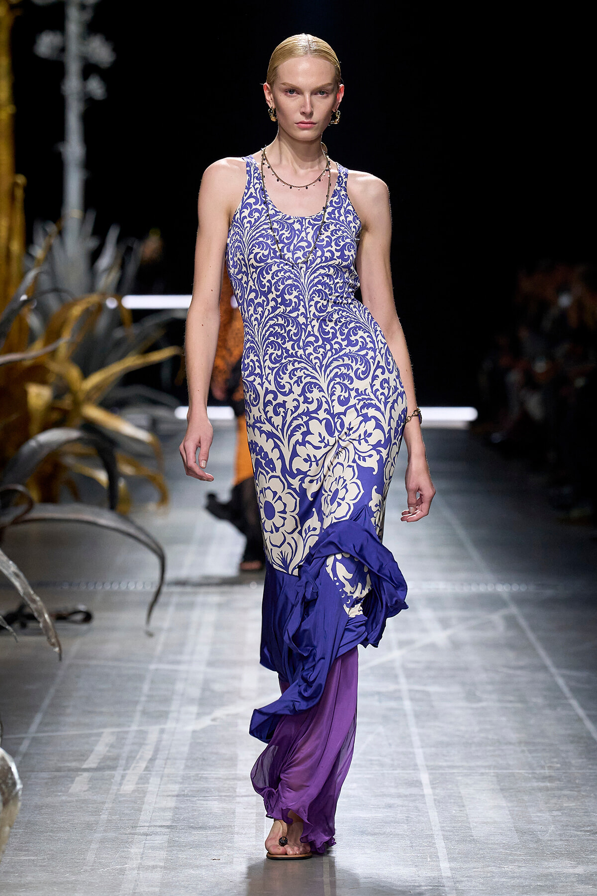 Model on a runway wearing a sleeveless blue and white damask dress with a purple ruffle hem and layered fabric at the bottom.