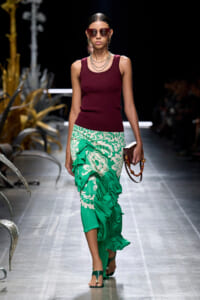 Model on a runway wearing a burgundy sleeveless top and a green floral-printed, gathered ruffled skirt, with oversized sunglasses and layered necklaces, carrying a small brown clutch.