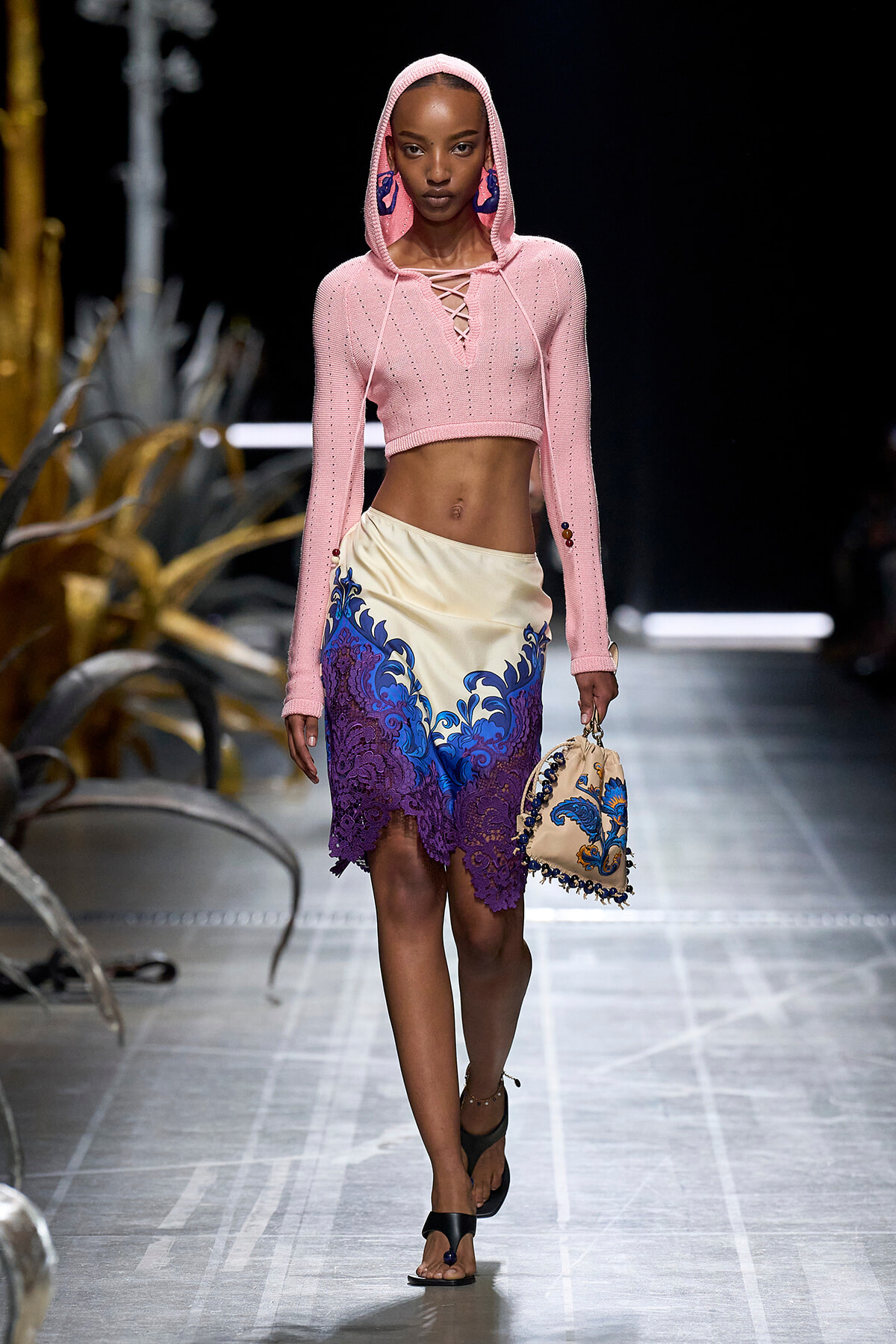 Model on a runway wearing a pink cropped hooded sweater with lace-up front and a cream skirt with blue-purple lace trim, carrying a beige embroidered handbag.