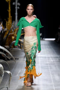 Model walks the runway in a bright green cropped knit top and a beige satin skirt with green and orange floral embroidery.