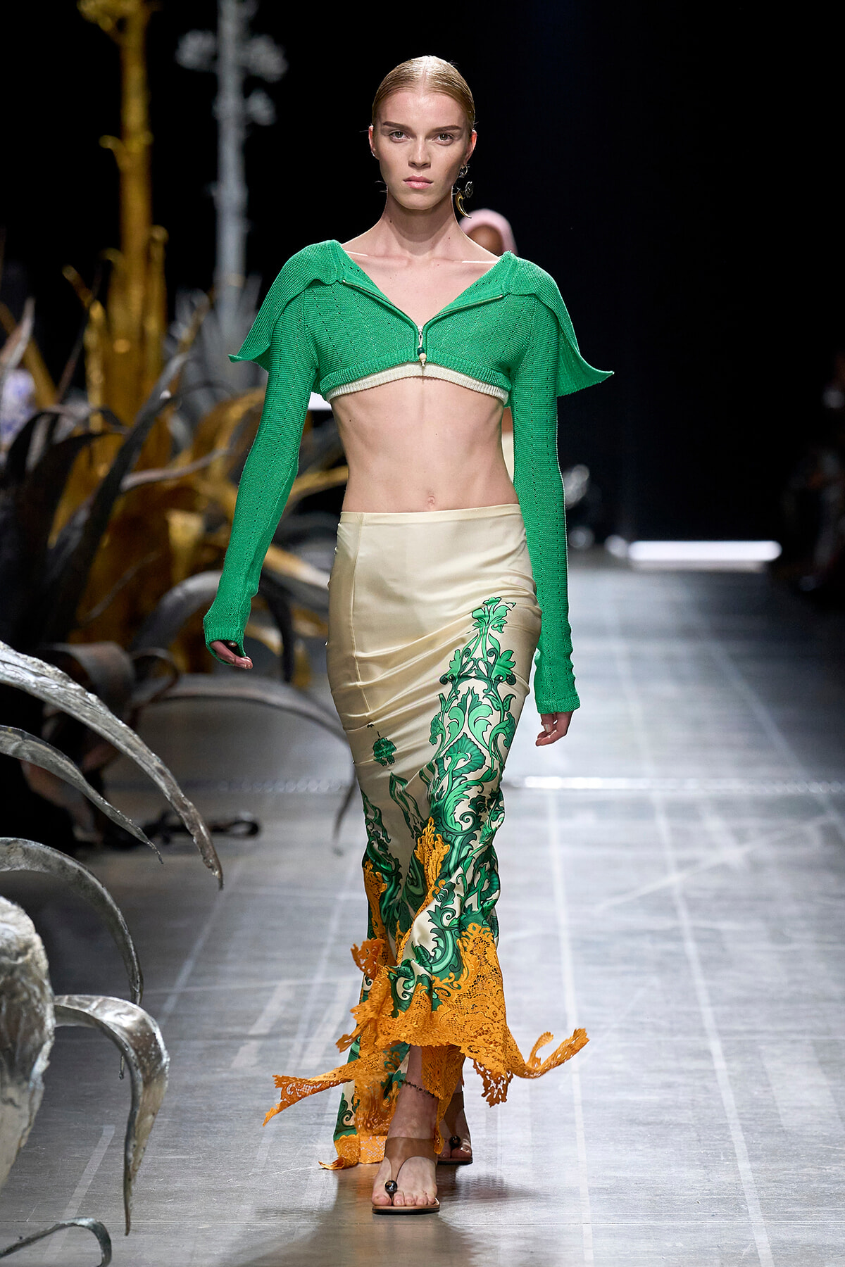Model walks the runway in a bright green cropped knit top and a beige satin skirt with green and orange floral embroidery.