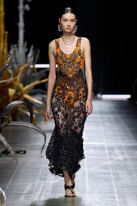 Model walks runway in an orange and black lace gown with intricate floral patterns and sheer black skirt, paired with bold earrings and sandals.
